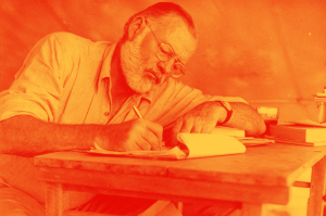 Foto de Hemingway, de óculos e barba branca, escrevendo à mão em uma mesa de madeira.