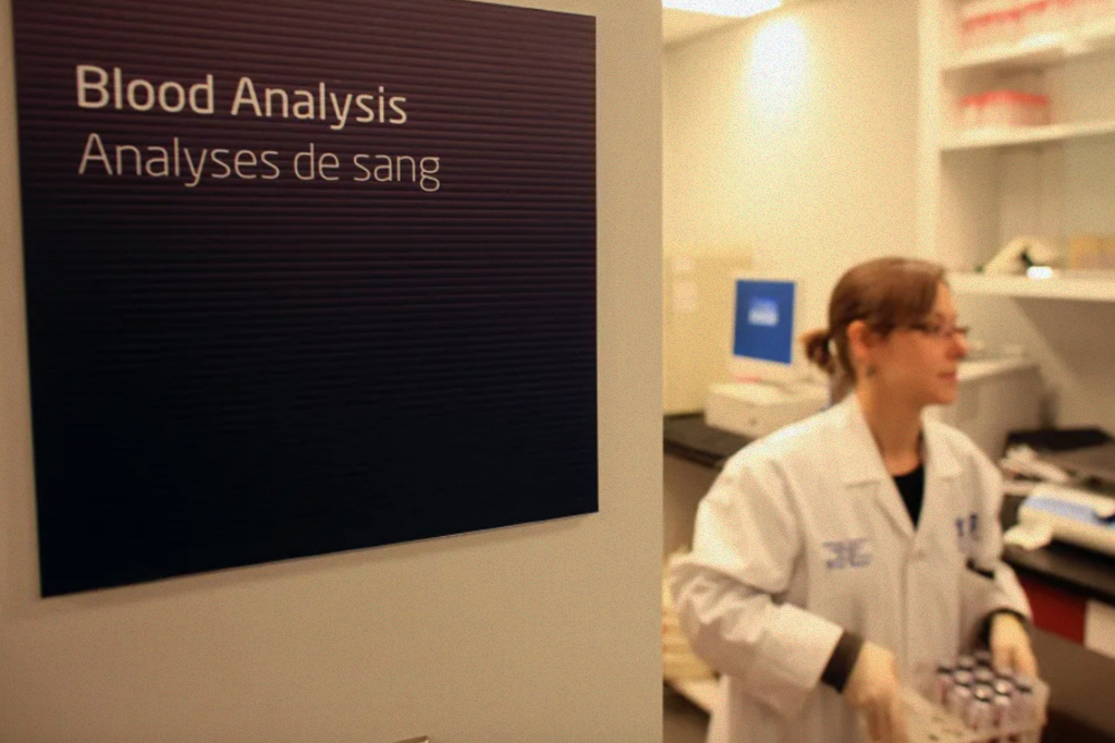 Foto de uma mulher saindo do laboratório de análise de sangue, carregando vários tubinhos de sangue.