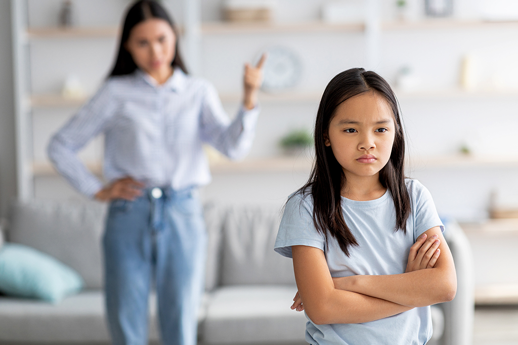 Mãe e filha brigando em casa. A menina está de cara fechada, ignorando sua mãe, de costas para ela com os braços cruzados.