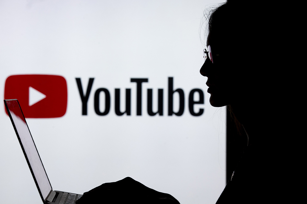 Silhueta de uma pessoa de perfil mexendo em um notebook, contra luz. No fundo, logo do Youtube.