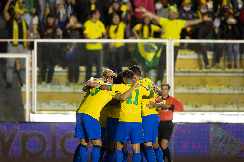 Fotografia de jogadores da seleção masculina principal se abraçando em campo, com torcedores comemorando ao fundo, desfocados.