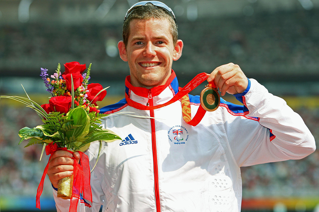 Retrato de John Mcfall segurando uma medalha de bronze no pódio.