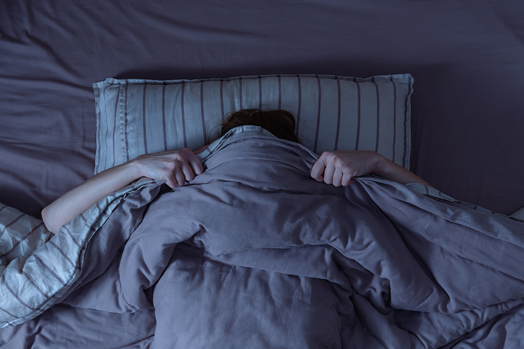 Pessoa deitada na cama se cobrindo -até o rosto- com edredom.