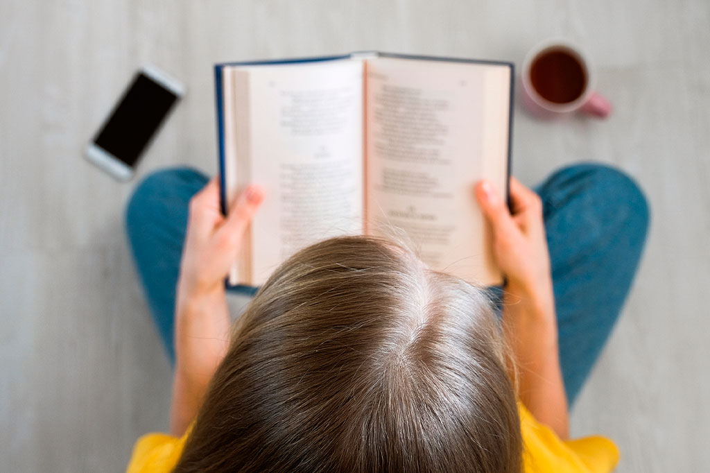 Visão superior de uma mulher sentada no chão com as pernas cruzadas na posição de lótus e lendo um livro. Ao lado está uma caneca de chá ou café e um celular.