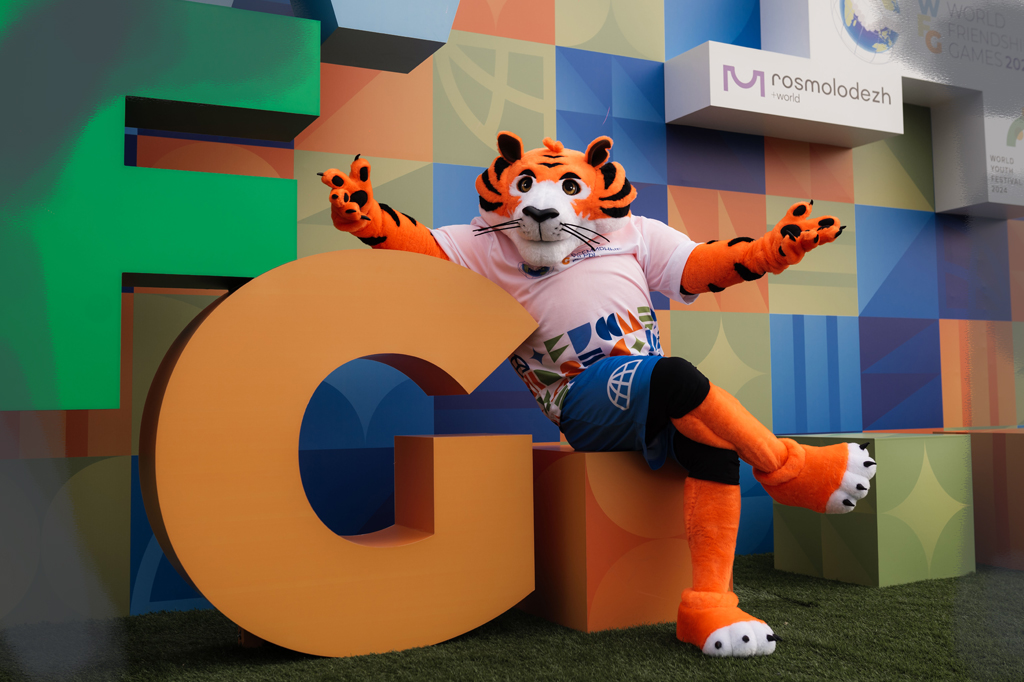 2606-jogos-amizade-russia-mascote-site Mascote de tigre em um cenário colorido.