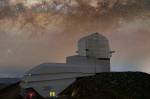 Observatório Rubin sob um céu noturno estrelado. O observatório fica no topo de seu cume rochoso em primeiro plano e é composto por um longo edifício branco que se estende para a esquerda e uma cúpula angular prateada projetando-se. A Via Láctea é visível, abrangendo a imagem horizontalmente acima do observatório como um denso fluxo de estrelas intercaladas com nuvens de poeira escuras e finas. O brilho amarelo das luzes da cidade em La Serena é visto ao longe, no canto inferior esquerdo.