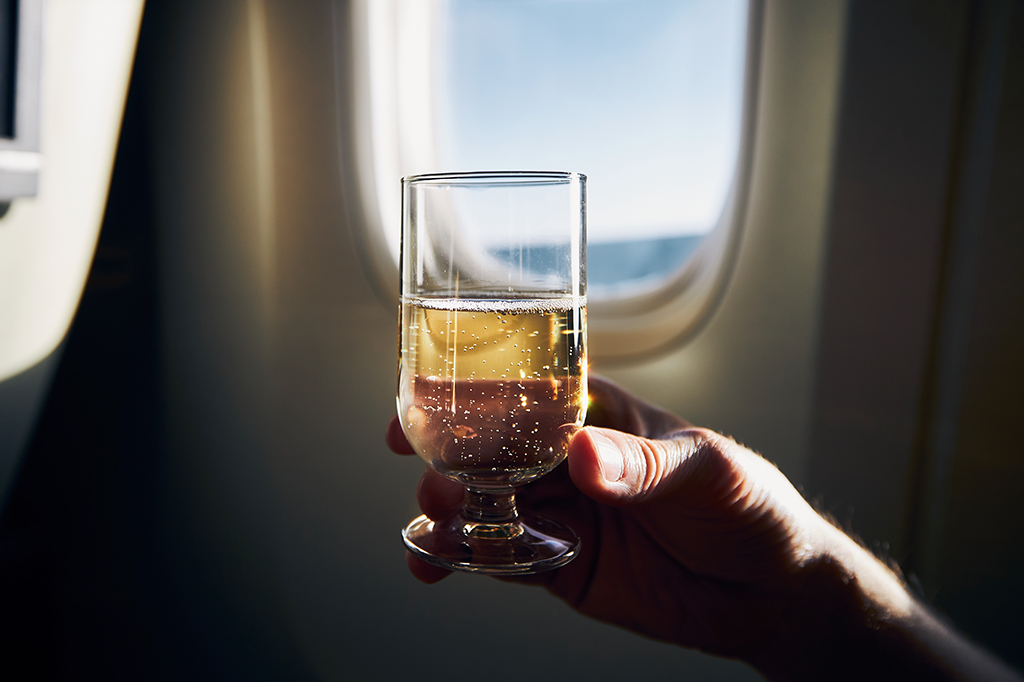 Uma mão segurando uma taça de espumante. Está de frente a uma janela de avião.