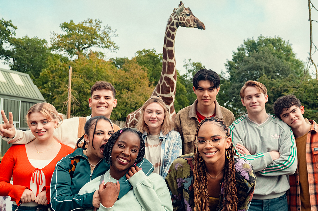 Grupo de jovens sorrindo para a foto. Ao fundo, há uma girafa e árvores.