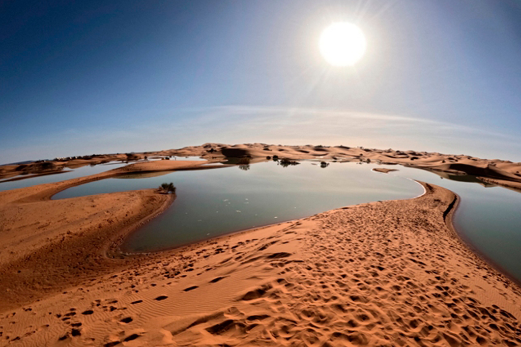 Foto do lago Erg Znaigui no deserto do Saara. Montes de areia em um dia com o céu limpo, com o sol raiando.