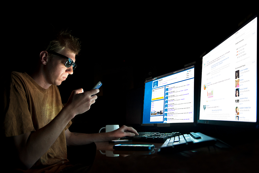 Imagem de um homem mexendo em dois computadores ao mesmo tempo e mexendo no celular.