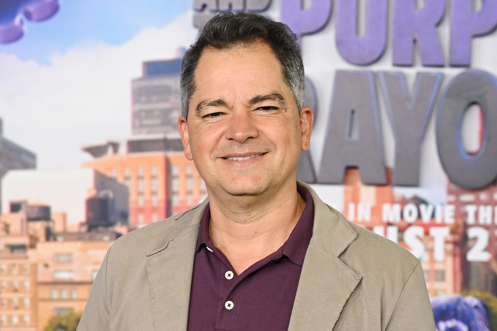Imagem de um homem sorridente, de cabelos grisalhos, vestindo terno marrom claro e camisa roxa.