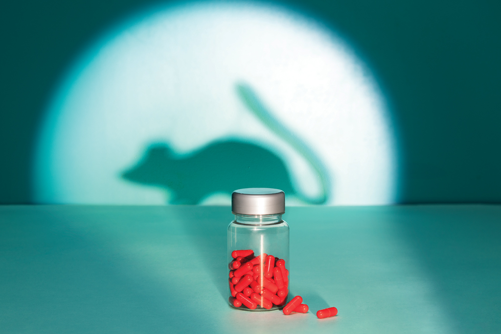 Imagem, de tom esverdeado, projetando a sombra de um rato na parede. Em primeiro plano, v&ecirc;-se, um frasco de vidro contendo c&aacute;psulas vermelhas.