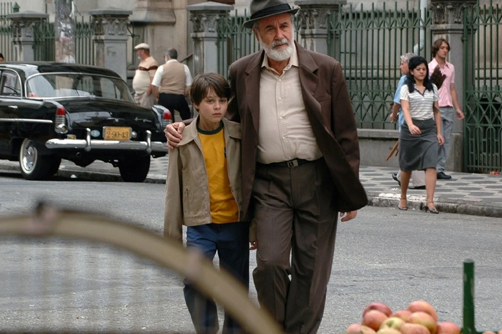 Imagem com cena do filme O Ano em que Meus Pais Sa&iacute;ram de F&eacute;rias (2006). V&ecirc;-se um garoto abra&ccedil;ado ao av&ocirc; caminhando pela rua.