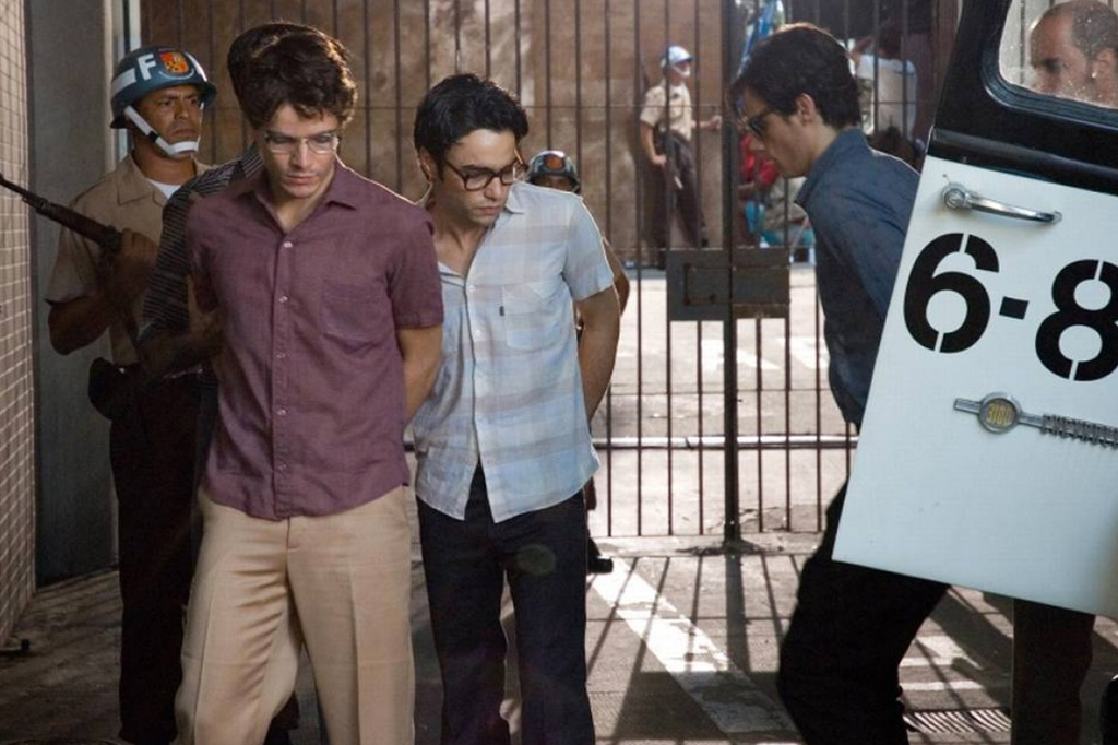 Imagem com cena do filme Batismo de Sangue (2007). V&ecirc;-se tr&ecirc;s jovens, com os bra&ccedil;os para tr&aacute;s, descendo de uma viatura policial.