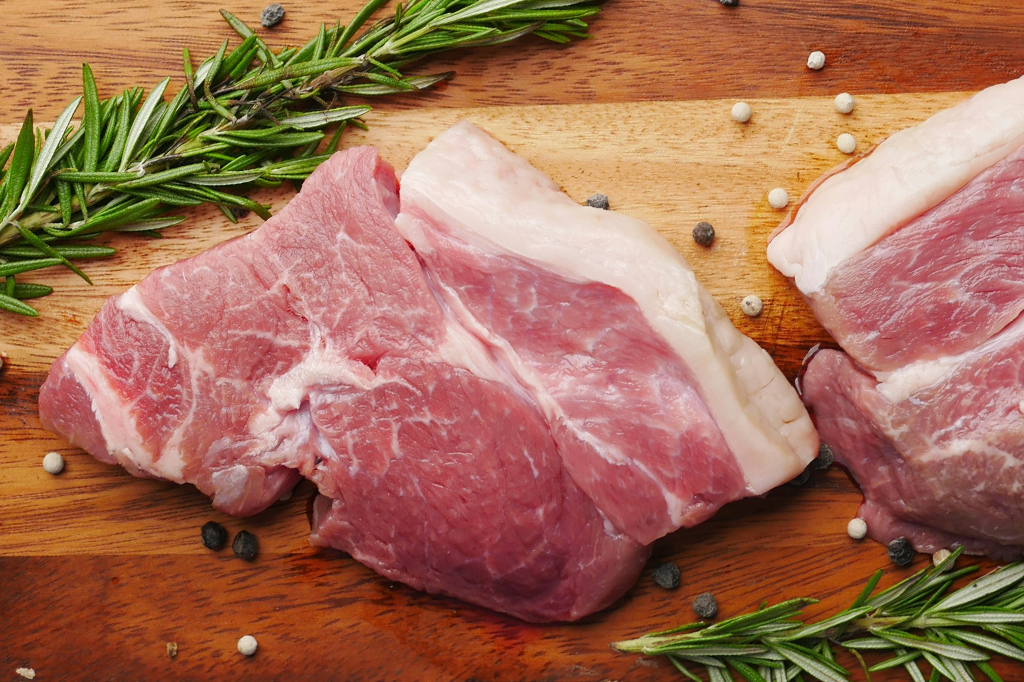 Imagem de dois pedaços de carne de porco em cima de uma tábua de madeira. Em volta, vê-se ramos de alecrim e bolinhas de pimenta do reino.