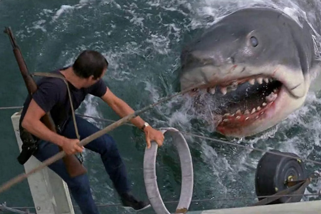 Imagem de um homem dependurado num barco enfrentando um enorme tubarão. Cena do filme "Tubarão".