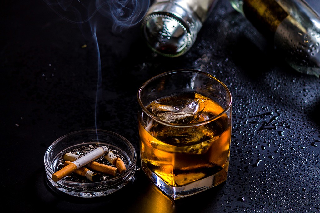 Copo de uísque com gelo e cinzeiro com cigarros acesos e bitucas sobre superfície escura molhada, com garrafas desfocadas ao fundo