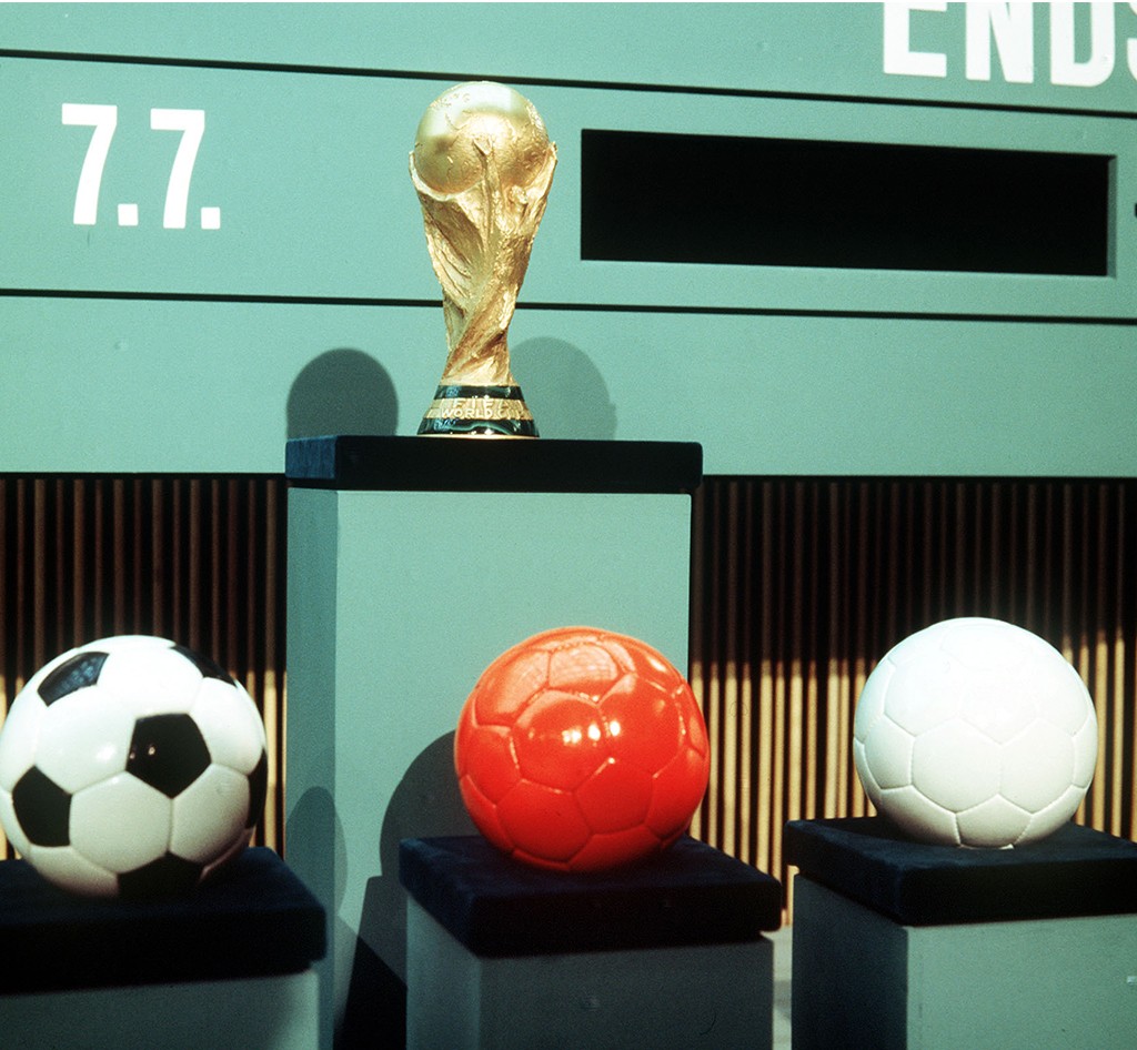 A Ta&ccedil;a da Copa do Mundo FIFA dourada em um pedestal preto, com tr&ecirc;s bolas de futebol em pedestais menores: uma branca e preta, uma laranja e uma branca. Ao fundo, um painel verde com a data 