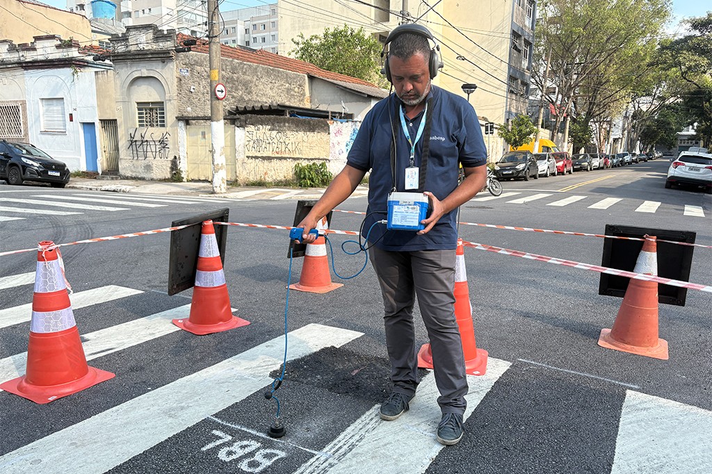 Homem de barba, camisa polo azul e fones de ouvido, usa equipamento para detectar vazamentos em rua com faixa de pedestres e cones de sinaliza&ccedil;&atilde;o