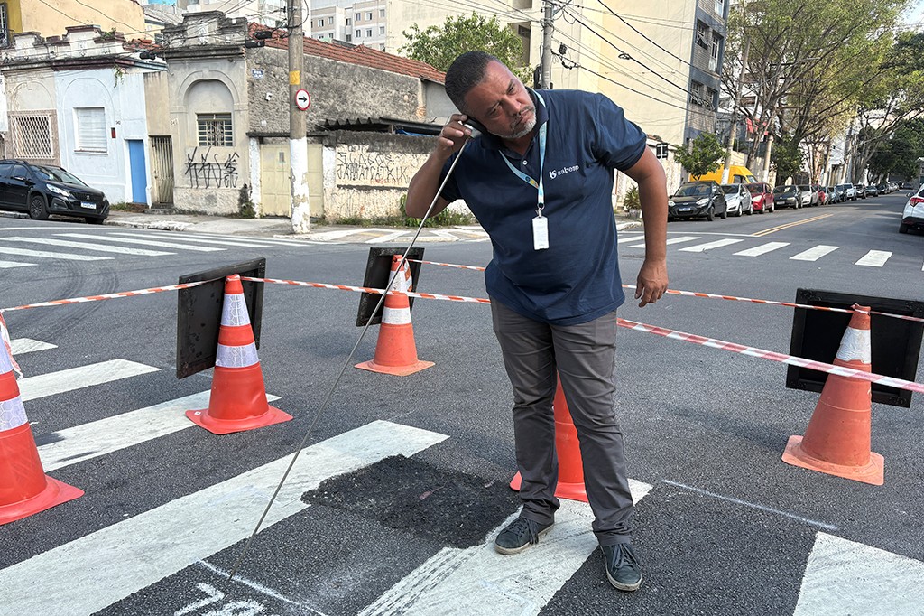 Homem de pele parda, barba e camiseta azul da Sabesp, inclina-se para ouvir um aparelho auditivo enquanto segura uma bengala longa. Ele est&aacute; em uma rua com faixa de pedestres, cones de sinaliza&ccedil;&atilde;o e fita zebrada, indicando obras. Ao fundo, carros estacionados e pr&eacute;dios antigos.