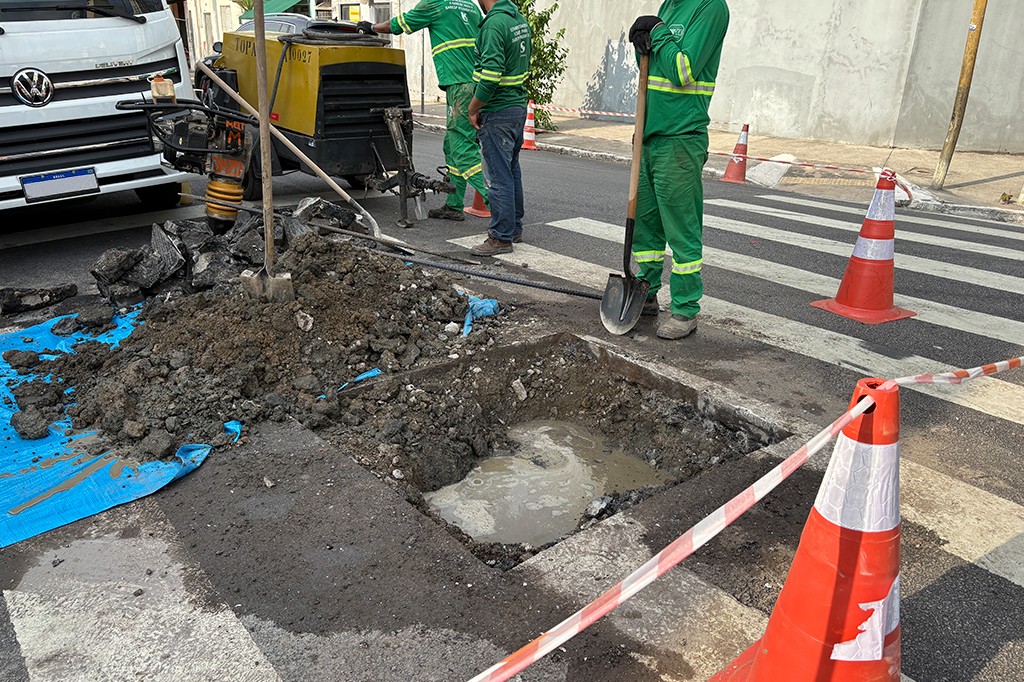 Tr&ecirc;s trabalhadores de uniforme verde operam equipamentos e uma p&aacute; em uma obra de rua, com um buraco cheio de &aacute;gua e terra, cones de sinaliza&ccedil;&atilde;o e uma faixa de pedestres ao fundo