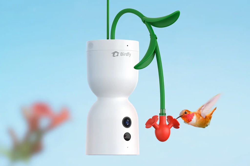C&acirc;mera de vigil&acirc;ncia Birdfy branca em formato de ampulheta, com um bra&ccedil;o verde em forma de folha e um alimentador vermelho de beija-flor, enquanto um beija-flor laranja se aproxima para beber n&eacute;ctar. Fundo azul claro.