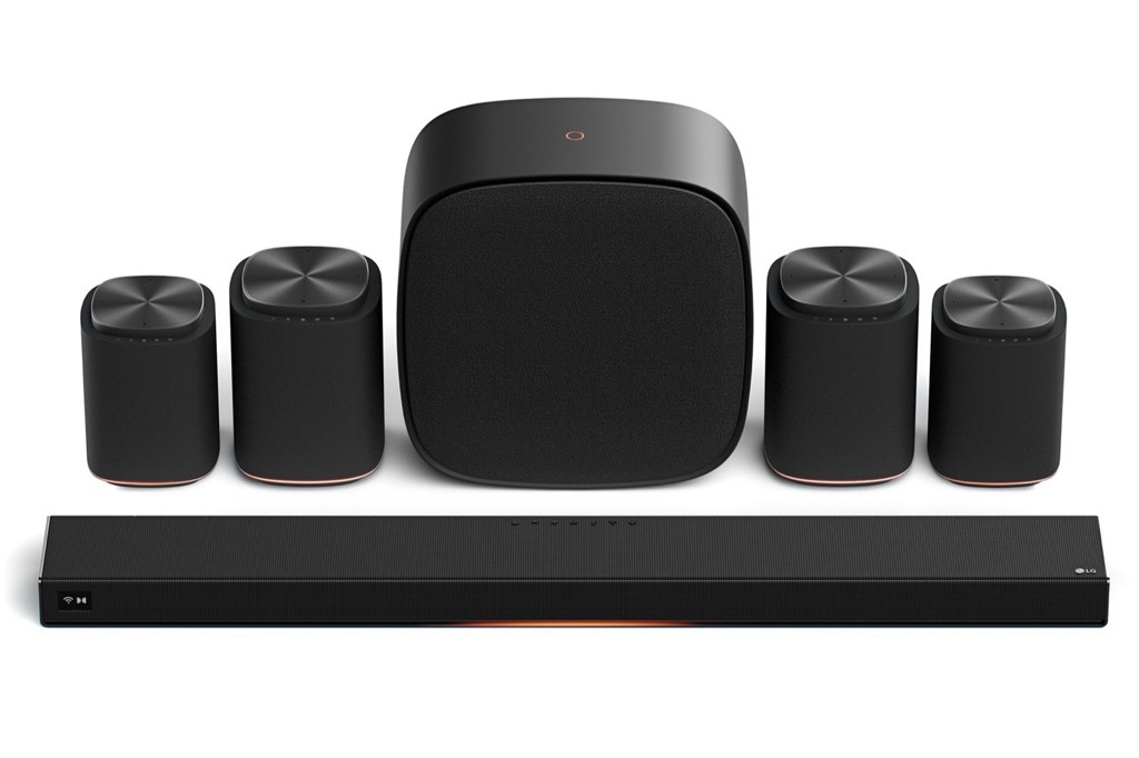 Sistema de som surround preto com soundbar retangular na frente, subwoofer central e quatro caixas de som cil&iacute;ndricas ao redor, todos com detalhes em cobre na base.