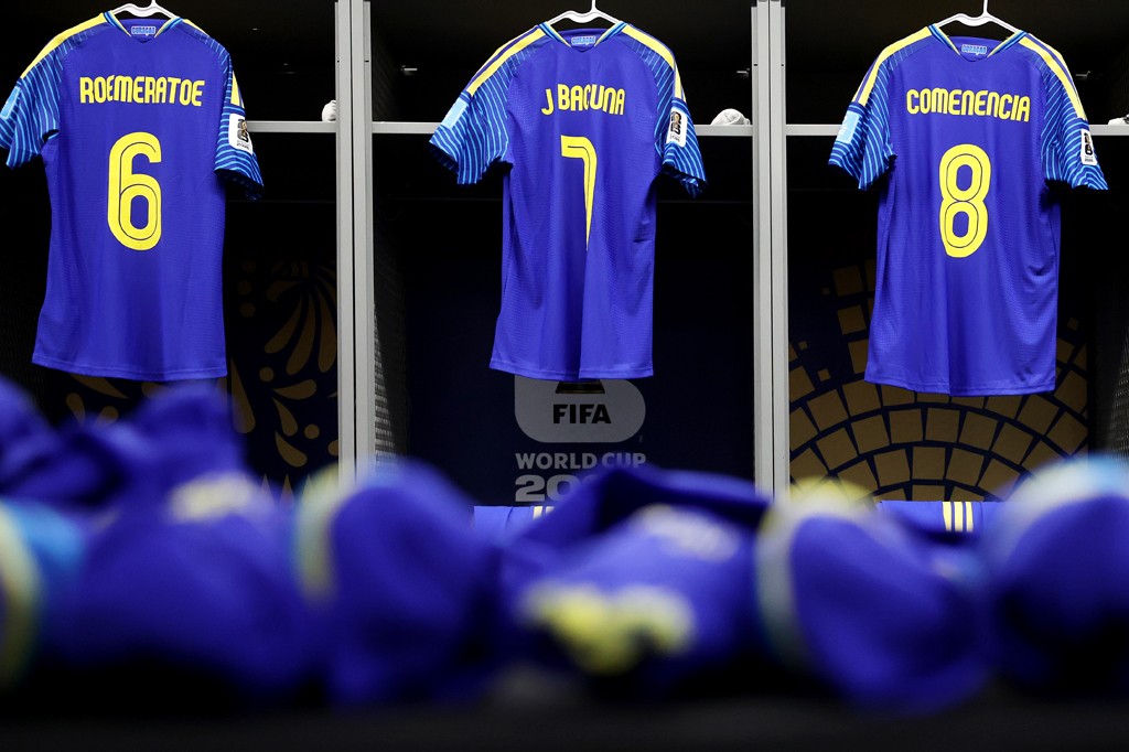 Três camisas de futebol da seleção de Curaçao, azuis com listras amarelas penduradas em armários de vestiário, com os nomes ROEMERATOE 6, J BACUNA 7 e COMENENCIA 8 em amarelo. No centro, um logo da FIFA World Cup 2022. Chuteiras azuis desfocadas em primeiro plano.