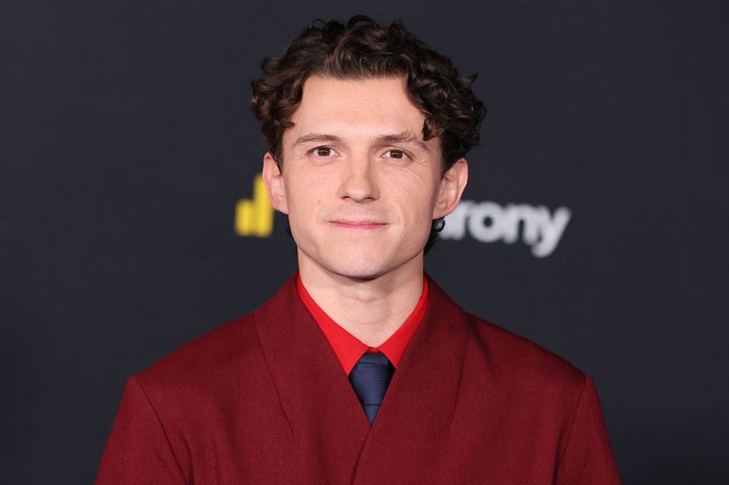 Tom Holland, jovem ator de cabelos castanhos encaracolados, sorri levemente para a c&acirc;mera. Ele veste um casaco vermelho-escuro sobre uma camisa vermelha e gravata azul-marinho. O fundo &eacute; escuro com um logo amarelo e branco desfocado.