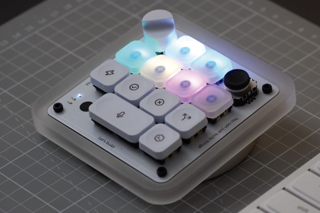 Um teclado modular branco com teclas iluminadas em tons de azul, roxo e branco, sobre uma base quadriculada cinza. Algumas teclas t&ecirc;m &iacute;cones como microfone, sigma e setas. H&aacute; um joystick preto no canto superior direito e texto Lets build e You can just build things na base