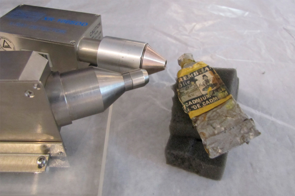 Equipamento de an&aacute;lise met&aacute;lico com dois bicos c&ocirc;nicos apontados para um tubo de tinta a &oacute;leo amassado, com r&oacute;tulo amarelado Rembrandt Olie e Cadmium sobre uma esponja cinza, em superf&iacute;cie branca