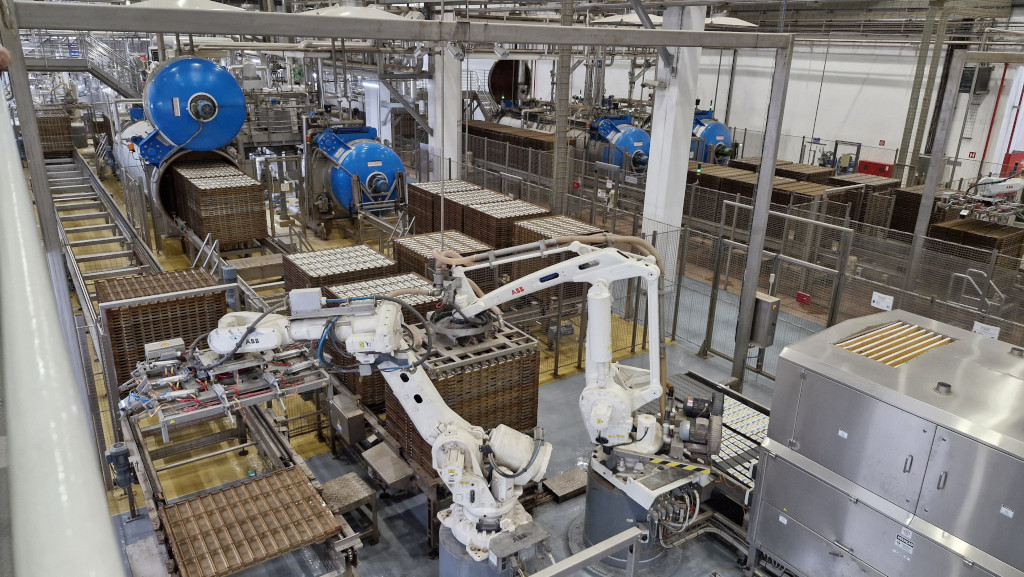 Vis&atilde;o a&eacute;rea de uma f&aacute;brica automatizada com esteiras transportadoras, rob&ocirc;s brancos da marca ABB e grandes m&aacute;quinas azuis cil&iacute;ndricas, processando caixas e bandejas de produtos em um ambiente industrial limpo