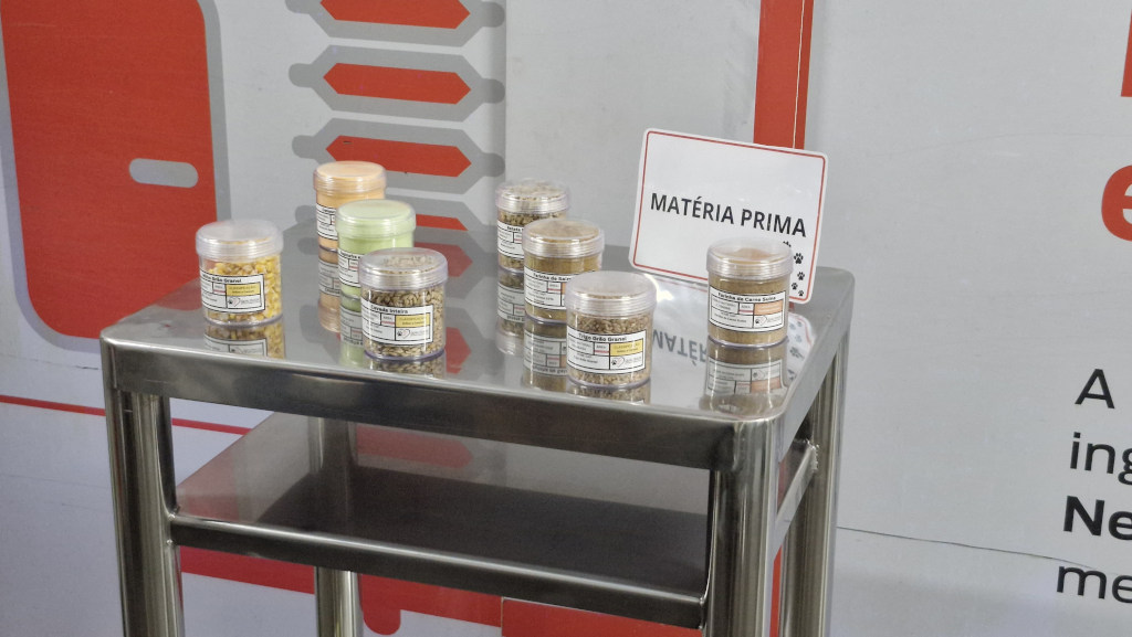 Nove potes transparentes com diferentes tipos de gr&atilde;os e farinhas, identificados com r&oacute;tulos brancos e amarelos, sobre uma mesa de metal. Ao fundo, um cartaz branco com a frase MAT&Eacute;RIA PRIMA em letras pretas e um desenho de patas de cachorro, e um painel branco com detalhes em vermelho e cinza
