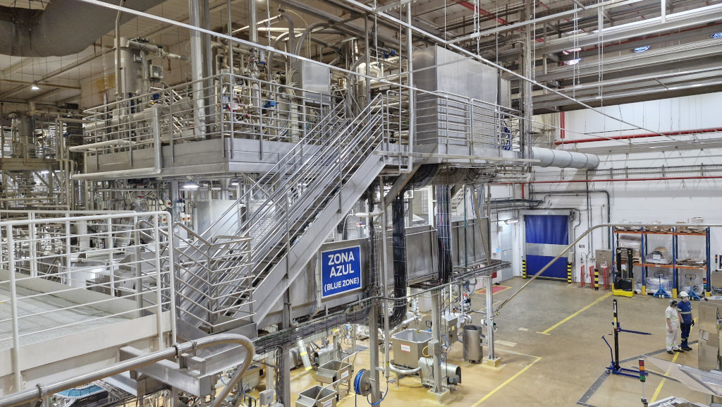 Interior de uma f&aacute;brica moderna com equipamentos industriais de a&ccedil;o inoxid&aacute;vel, tubula&ccedil;&otilde;es e plataformas elevadas. Uma escada met&aacute;lica central leva a um n&iacute;vel superior. Um letreiro azul na estrutura indica ZONA AZUL (BLUE ZONE). No ch&atilde;o, dois trabalhadores com capacetes e uniformes brancos e azuis observam o ambiente. O piso &eacute; amarelo com faixas de seguran&ccedil;a, e ao fundo h&aacute; uma porta azul e prateleiras com materiais