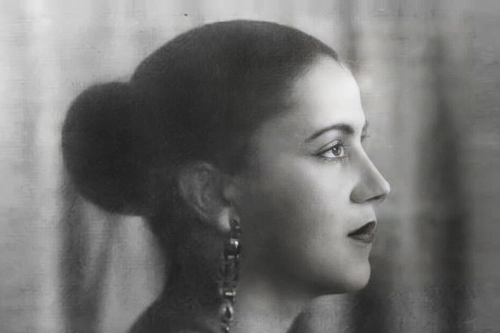 Retrato em preto e branco de uma mulher jovem, de perfil, olhando para a direita. Ela tem cabelo escuro preso em um coque baixo, maquiagem nos olhos e lábios, e um brinco longo e escuro pendurado na orelha esquerda. A expressão é serena, com a boca levemente aberta